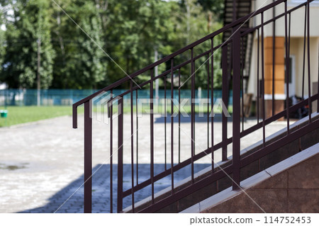 Close-up outdoors detail of short stone staircase with metal protective simple primitive metal fence and handrails on lit by bright summer sun big yard and blurred tall green trees background. 114752453