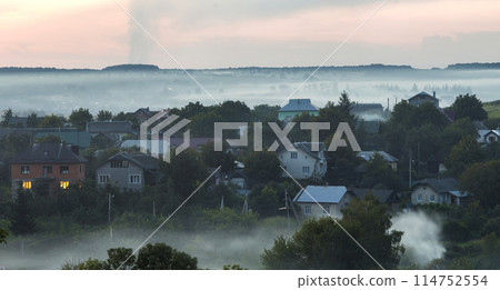 Beautiful peaceful top view summer countryside panorama at sunrise. Residential cottages among green trees covered with blue fog, distant woody horizon and light pink sky. Beauty of nature concept. Beautiful peaceful top view summer countryside panorama at sunrise. Residential cottages among green trees covered with blue fog, distant woody horizon and light pink sky. Beauty of nature concept. 114752554