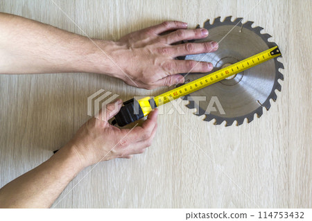 Close up of circular saw blade for wood work and worker hand with measuring tape on wooden background. Top view. Close up of circular saw blade for wood work and worker hand with measuring tape on wooden background. Top view. 114753432
