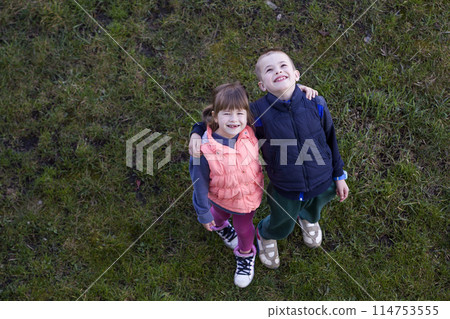 View from above of two cute children in casual clothes, boy and girl standing on green grass, hugging each other over shoulders, looking up and happily smiling. Happy childhood and friendship concept. 114753555