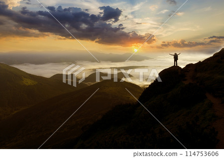 Wide mountain panorama. Small silhouette of tourist with backpack on rocky mountain slope with raised hands over valley covered with white puffy clouds. Beauty of nature, tourism and traveling concept 114753606