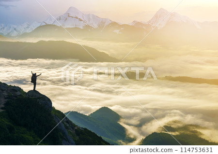 Wide mountain panorama. Small silhouette of tourist with backpack on rocky mountain slope with raised hands over valley covered with white puffy clouds. Beauty of nature, tourism and traveling concept 114753631