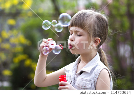 Profile of pretty small preschool blond girl with funny serious expression blowing colorful transparent soap bubbles outdoors on blurred green summer background. Joy of careless childhood concept. Profile of pretty small preschool blond girl with funny serious expression blowing colorful transparent soap bubbles outdoors on blurred green summer background. Joy of careless childhood concept. 114753654
