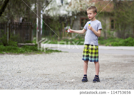 Young handsome blond schoolboy in casual clothing with funny serious expression blows transparent soap bulbs standing outdoors on warm summer day on rural background. Careless happy childhood concept. Young handsome blond schoolboy in casual clothing with funny serious expression blows transparent soap bulbs standing outdoors on warm summer day on rural background. Careless happy childhood concept. 114753656