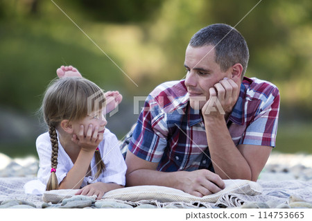 Portrait of young father and cute blond girl with long braids laying on pebbled river bank on bright summer day on blurred green trees bokeh background. Love, care and perfect holiday concept. 114753665