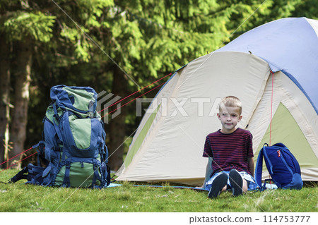 Holiday camping. Young boy sitting in front of a tent near backpacks taking rest after hiking in the forest shows something in the trees. Traveling and outdoors activities. Holiday camping. Young boy sitting in front of a tent near backpacks taking rest after hiking in the forest shows something in the trees. Traveling and outdoors activities. 114753777