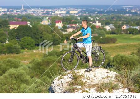 Young man with a bicycle on a rock looking on a city Young man with a bicycle on a rock looking on a city 114754085