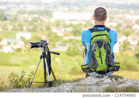 Hiker photographer sitting on a rock looking forward near a camera on a tripod 114754093