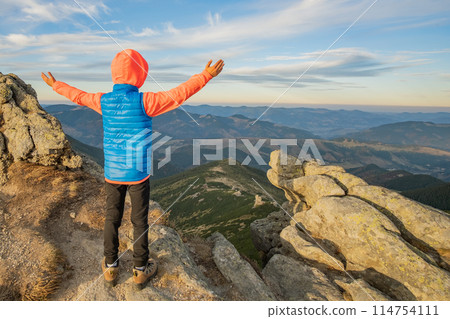 Young child boy hiker standing with raised hands in mountains enjoying view of amazing mountain landscape at sunset. 114754111