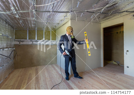 Strange man in businessman suit and gas protection mask inside a room under renovation works holding electric screwdriver and a level tools. Strange man in businessman suit and gas protection mask inside a room under renovation works holding electric screwdriver and a level tools. 114754177