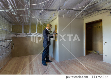 Strange man in businessman suit and gas protection mask inside a room under renovation works holding electric screwdriver and a level tools. 114754186
