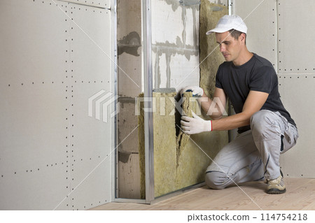 Worker in protective gloves insulating rock wool insulation in wooden frame for future house walls for cold barrier. Comfortable warm home, economy, construction and renovation concept Worker in protective gloves insulating rock wool insulation in wooden frame for future house walls for cold barrier. Comfortable warm home, economy, construction and renovation concept 114754218