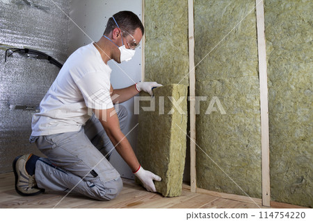 Worker in protective goggles and respirator insulating rock wool insulation in wooden frame for future house walls for cold barrier. Comfortable warm home, economy, construction and renovation concept Worker in protective goggles and respirator insulating rock wool insulation in wooden frame for future house walls for cold barrier. Comfortable warm home, economy, construction and renovation concept 114754220