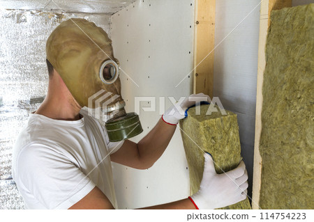 Young worker in gas mask insulating rock wool insulation staff in wooden frame for future house walls for cold and heat barrier. Comfortable warm home, economy, construction and renovation concept. 114754223
