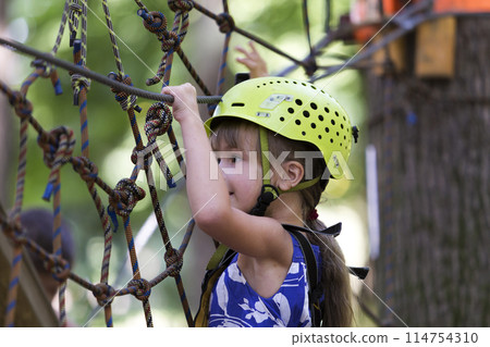 Young pretty blond child girl in safety harness and helmet on rope way on bright bokeh background. 114754310