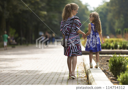 Back view of young blond long-haired attractive woman and small child girl holding hands outdoors. 114754312