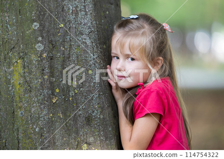 Portrait of a pretty little child girl standing near big tree trunk in summer park outdoors. Portrait of a pretty little child girl standing near big tree trunk in summer park outdoors. 114754322
