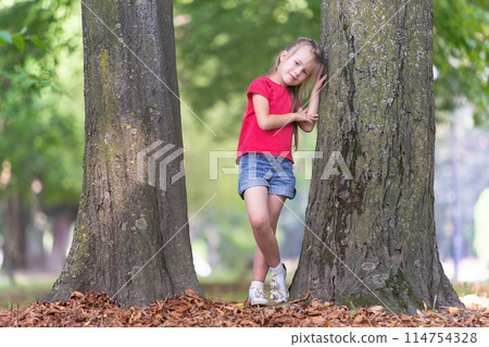 Portrait of a pretty little child girl standing near big tree trunk in summer park outdoors. 114754328