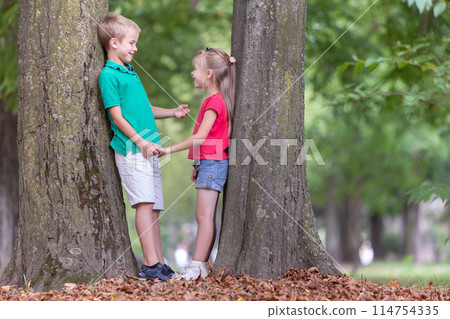 Portrait of two pretty cute children boy and girl standing near big tree trunk in summer park outdoors. 114754335