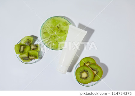 Minimalist style photograph of kiwi ingredient cosmetic theme, a blank tube in white color placed in center with a few glass dishes of fresh kiwi around, over white background. Mock up for design 114754375