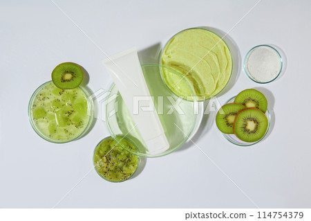 Photograph for exfoliating cream has kiwi ingredient advertising , a blank scrubs tube displayed on a glass dish with a little light green liquid, white background. High angle shot, space for text 114754379