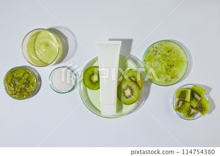 Flat lay glassware containing fresh kiwi, green essence and yellow exfoliating scrubs above, green color of kiwi striking on white countertop. Unlabeled cosmetic tube in center, space for text Flat lay glassware containing fresh kiwi, green essence and yellow exfoliating scrubs above, green color of kiwi striking on white countertop. Unlabeled cosmetic tube in center, space for text 114754380