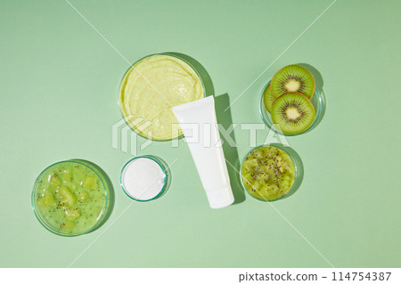 Flat lay kiwi ingredient contained on glass dishes over pastel green texture, a exfoliating cream tube without label leaning on a glass dish of yellow exfoliation. Space for designing 114754387