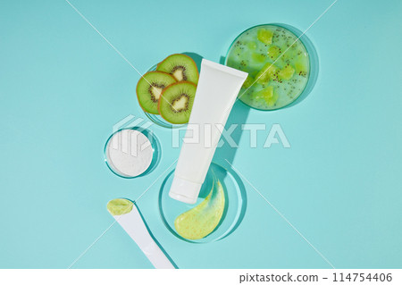 Creative photo for natural exfoliation products, blue countertop containing some glassware in round shaped with fresh kiwi, a white tube without label featured on top. High angle shot, vacant space 114754406