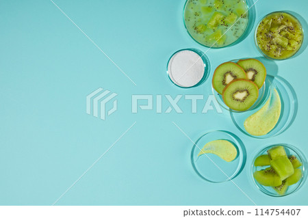 Top-down photo of a blue table top with lots of round glass utensils containing sliced, chopped kiwi, yellow and white exfoliating creams. Copy space, blank on the left side Top-down photo of a blue table top with lots of round glass utensils containing sliced, chopped kiwi, yellow and white exfoliating creams. Copy space, blank on the left side 114754407