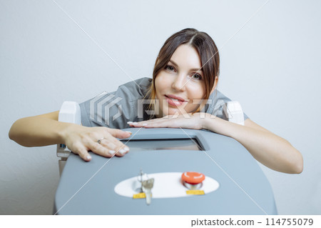 Female cosmetologist in a medical coat holds a working part of the modern laser epilator in her hands and poses for a photo. Laser epilation treatment in cosmetic beauty clinic. 114755079