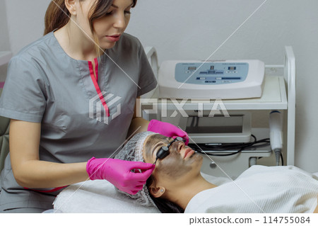 Close-up of carbon peeling procedure for middle-aged woman face. Laser pulses clean skin. Process of photothermolysis, warming, laser carbon peeling. Facial skin rejuvenation, cosmetology treatment. Close-up of carbon peeling procedure for middle-aged woman face. Laser pulses clean skin. Process of photothermolysis, warming, laser carbon peeling. Facial skin rejuvenation, cosmetology treatment. 114755084