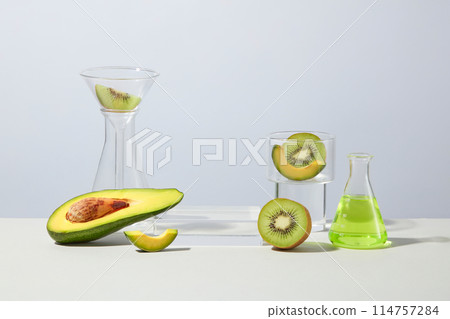 A group of laboratory glassware and natural ingredient like avocado and kiwi featured on light blue background with an empty pedestal in center for showing product. Blank space for design, front view 114757284