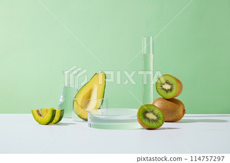Green background template of avocado and kiwi theme with laboratory concept, empty round glass platform, fresh kiwi and avocado, measuring cylinder and erlenmeyer flask. Copy space, front view 114757297