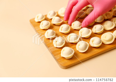 A chef's hand in a rubber glove carefully lays out semi-cooked raw dumplings on a wooden board, copy space. 114758782