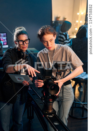 Vertical portrait of camerawoman operating professional video camera on rig with female director giving instruction Vertical portrait of camerawoman operating professional video camera on rig with female director giving instruction 114758882