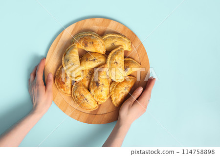 Top view on woman hands with wooden plate with baked samsa pies on blue background. Top view on woman hands with wooden plate with baked samsa pies on blue background. 114758898