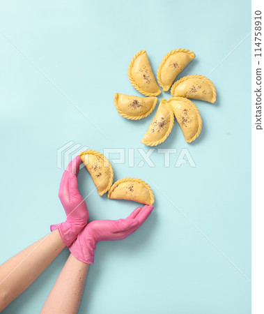 Top view a woman baker's hands in a rubber gloves lays out raw samsa on blue background. Traditional indian cuisine, frozen semi-finished products. Top view a woman baker's hands in a rubber gloves lays out raw samsa on blue background. Traditional indian cuisine, frozen semi-finished products. 114758910
