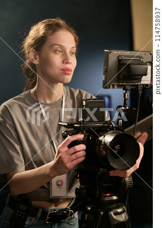 Vertical portrait of smiling Caucasian camerawoman operating equipment on set during video production 114758937
