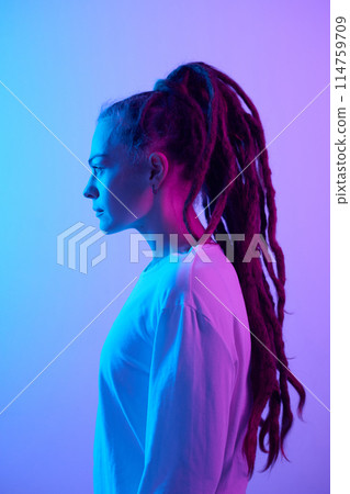 Vertical side view portrait of Caucasian young woman with long dreadlocks hairstyle standing in blue and purple neon light Vertical side view portrait of Caucasian young woman with long dreadlocks hairstyle standing in blue and purple neon light 114759709