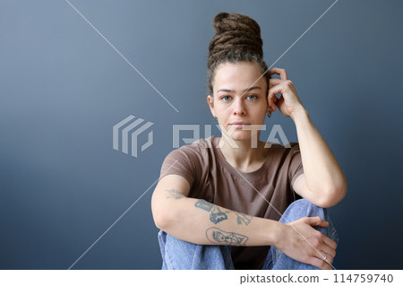 Minimal portrait of tattooed Caucasian woman with dreadlocks in bun hairstyle looking at camera sitting on grey background copy space Minimal portrait of tattooed Caucasian woman with dreadlocks in bun hairstyle looking at camera sitting on grey background copy space 114759740