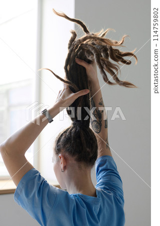 Vertical back view of young Caucasian woman holding long dreadlocks in ponytail facing window Vertical back view of young Caucasian woman holding long dreadlocks in ponytail facing window 114759802