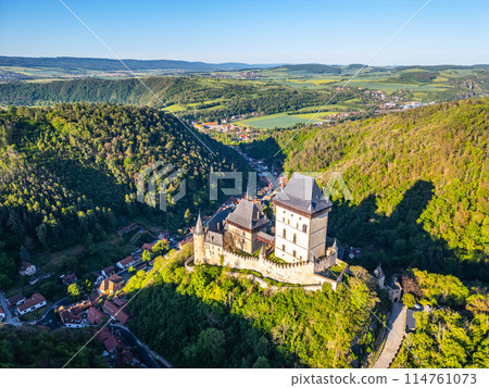 Aerial perspective of Karlstejn Castle nestled in a forested landscape with surrounding lush greenery on a clear, sunny day in Czechia. Aerial view from drone. Aerial perspective of Karlstejn Castle nestled in a forested landscape with surrounding lush greenery on a clear, sunny day in Czechia. Aerial view from drone. 114761073