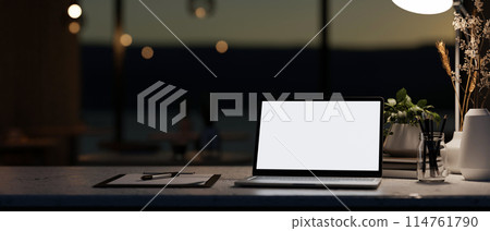 A modern dark office at night featuring a white-screen laptop computer mockup on a table. 114761790