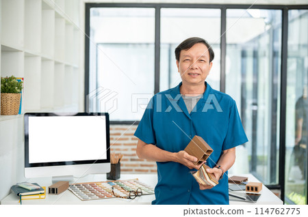 A senior Asian male interior designer with material samples in his hand is standing in his office. 114762775