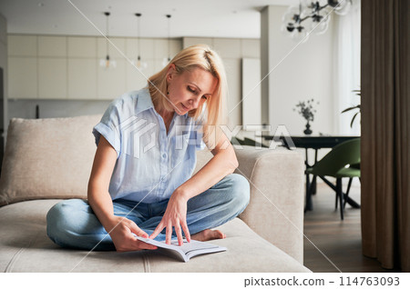 Woman sitting comfortably on couch, deeply engaged in reading book. Cozy living space with modern aesthetic on background. Person enjoying quiet moment of solitude, immersed in reading. 114763093