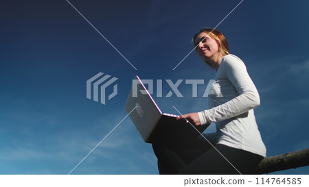 Young woman in modern clothing sits gracefully on a rustic wooden fence, absorbed in her laptop screen. The sun shines down on her as she navigates the digital world, blending nature and technology. Young woman in modern clothing sits gracefully on a rustic wooden fence, absorbed in her laptop screen. The sun shines down on her as she navigates the digital world, blending nature and technology. 114764585