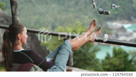 Woman sitting on a bench with feet propped up. Female relax outdoor on terrace with sea view. Tourist girl barefoot enjoy outdoor lifestyle travel on summer holiday vacation. Relaxation, silence Woman sitting on a bench with feet propped up. Female relax outdoor on terrace with sea view. Tourist girl barefoot enjoy outdoor lifestyle travel on summer holiday vacation. Relaxation, silence 114764588