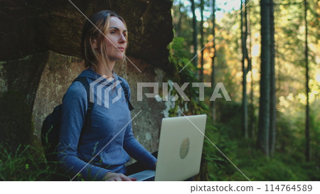 A young woman dressed in earthy tones is sitting cross-legged on mossy ground in a mystical forest, typing on a laptop with rays of sunlight filtering through the canopy above her. 114764589