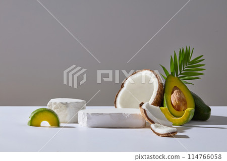White countertop containing an empty plaster pedestals, dry coconut and fresh avocado, decorated by a tree branch over light gray background. Frontal shot photo, space for showing product 114766058