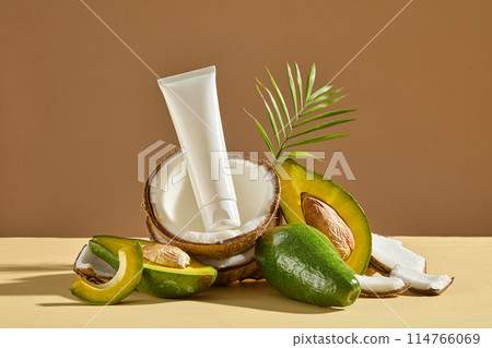 Coconuts and avocados are arranged in a small pile on light yellow countertop with an unlabeled white cosmetics tube displayed on top over brown background. Mock up for design with copy space 114766069